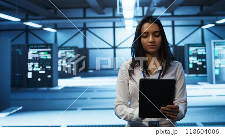 Software developer walking in data center, using tablet to design, implement and manage gear, ensuring optimal performance and reliability. IT worker in server farm coding on device, camera B 118604006