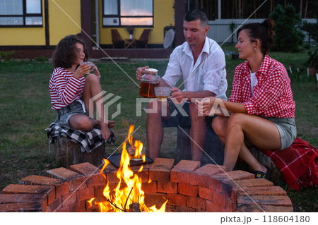 Family mom, dad, daughter are drinking tea in the courtyard of the house by the fire pit on a cozy summer evening. Family mom, dad, daughter are drinking tea in the courtyard of the house by the fire pit on a cozy summer evening. 118604180