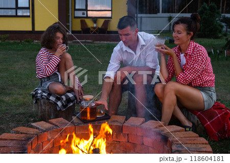 Family mom, dad, daughter are drinking tea in the courtyard of the house by the fire pit on a cozy summer evening. Family mom, dad, daughter are drinking tea in the courtyard of the house by the fire pit on a cozy summer evening. 118604181