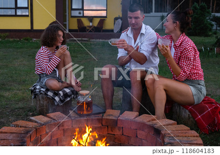 Family mom, dad, daughter are drinking tea in the courtyard of the house by the fire pit on a cozy summer evening. Family mom, dad, daughter are drinking tea in the courtyard of the house by the fire pit on a cozy summer evening. 118604183