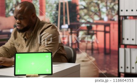 Businessman examines annual sales reports on documents next to a green screen tablet display, doing an accounting analytics plan. Entrepreneur working remotely from home office. Camera A. Businessman examines annual sales reports on documents next to a green screen tablet display, doing an accounting analytics plan. Entrepreneur working remotely from home office. Camera A. 118604353