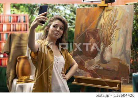 In a modern atelier, artist is filming her rustic artwork painting with the smartphone, social media branding. Recording and taking pictures of colorful canvas with watercolor piece, self promotion. 118604426