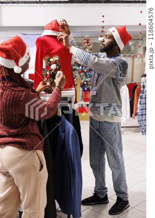 Mall employees decorating clothing store for christmas holiday season, using wreaths, santa hats and balls to create festive atmosphere in boutique. African american people working on xmas decor. Mall employees decorating clothing store for christmas holiday season, using wreaths, santa hats and balls to create festive atmosphere in boutique. African american people working on xmas decor. 118604576