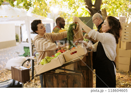 Female food delivery worker taking bag of veggies to give order to clients. Couple of local farmers standing behind fruit and vegetable stand at farmers market, giving package to deliverywoman. Female food delivery worker taking bag of veggies to give order to clients. Couple of local farmers standing behind fruit and vegetable stand at farmers market, giving package to deliverywoman. 118604605