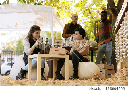 Shopping at an organic food festival, a young woman sits at a table with a local winemaker and tastes homemade natural wine before making a purchase. People sampling products at the farmers market. 118604606