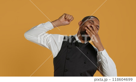 Tired waiter yawning and stretching after a night shift and feeling exhausted, sleepy employee falling asleep in studio. Hospitality industry staff works in customer service, burnout. Camera A. Tired waiter yawning and stretching after a night shift and feeling exhausted, sleepy employee falling asleep in studio. Hospitality industry staff works in customer service, burnout. Camera A. 118604699