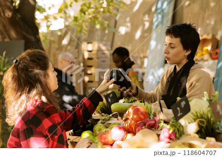 Vendor woman talking to female customer and selling fresh locally grown produce at farmers market stand. Seller helping client to choose veggies, explaining health benefits of organic food. Vendor woman talking to female customer and selling fresh locally grown produce at farmers market stand. Seller helping client to choose veggies, explaining health benefits of organic food. 118604707