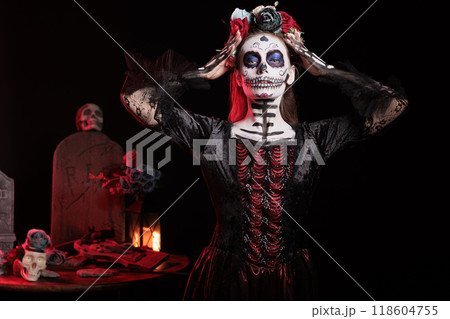 Portrait of person at religious processions during Dia de los Muertos november holiday. Woman wearing skull body paint and floral crown symbolizing reverence for the deceased, dark studio background Portrait of person at religious processions during Dia de los Muertos november holiday. Woman wearing skull body paint and floral crown symbolizing reverence for the deceased, dark studio background 118604755