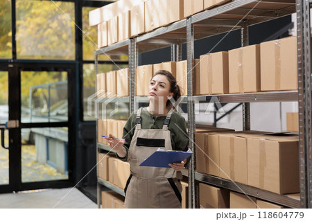 Storage room worker searching clients parcel in storehouse, analyzing clipboard with online order. Warehouse manager wearing uniform overalls supervising merchandise in factory storehouse 118604779