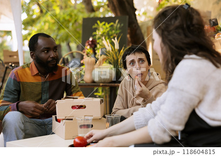 Caucasian farmer offering freshly harvested bio produce to multiracial customers. Young diverse consumers seated and tasting locally grown organic fruits at harvest farm fair festival. Caucasian farmer offering freshly harvested bio produce to multiracial customers. Young diverse consumers seated and tasting locally grown organic fruits at harvest farm fair festival. 118604815