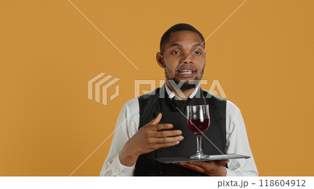 Waiter serving clients with a glass of red wine before dinner, recommending a special drink for customer satisfaction. Employee carrying a tray and serves people with professionalism. Camera A. Waiter serving clients with a glass of red wine before dinner, recommending a special drink for customer satisfaction. Employee carrying a tray and serves people with professionalism. Camera A. 118604912