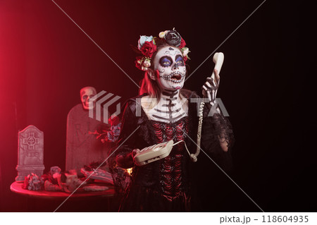 Dark lady talking on office cord telephone during Dia de los Muertos, preserving cultural traditions. Woman using landline phone to answer call, wearing la Catrina costume to celebrate Mexican culture 118604935