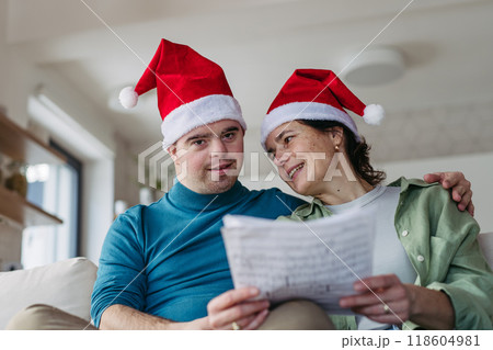 Young man with Down syndrome learning and singing christmas songs with mother. Concept of peaceful Christmas family moment. Young man with Down syndrome learning and singing christmas songs with mother. Concept of peaceful Christmas family moment. 118604981