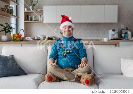 Portrait of young man with Down syndrome with santa's hat on head and christmas light around body, looking at camera. Portrait of young man with Down syndrome with santa's hat on head and christmas light around body, looking at camera. 118605002