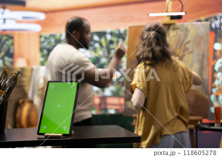 Two artists painting on a canvas next to green screen on tablet display, working together on their new masterpiece for fine art gallery exhibition. Diverse team of painters in art studio. 118605278