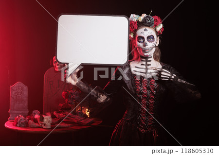 Portrait of woman dressed in Day of the Dead costume celebrating Dia de los Muertos, holding speech bubble. Girl wearing skeleton makeup using isolated copyspace template, promoting Mexican culture 118605310