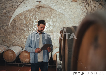 The winemaker standing in the wine cellar, monitoring the fermentation and aging processes in large barrels. The winemaker standing in the wine cellar, monitoring the fermentation and aging processes in large barrels. 118606139