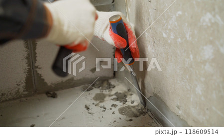 Floor renovation concept. Cleaning the walls from excess hardened mortar after dismantling with a flat chisel and removing debris. The master with the help of a chisel and a hammer removes the Floor renovation concept. Cleaning the walls from excess hardened mortar after dismantling with a flat chisel and removing debris. The master with the help of a chisel and a hammer removes the 118609514