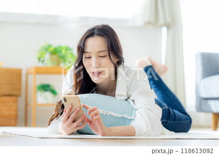 リビングのテーブルで寝転んでスマホを見る若い女性 118613092