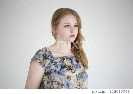 Young woman with long hair poses thoughtfully in a floral blouse against a plain background. The neutral background highlights her thoughtful demeanor. Young adult, european. 118613798