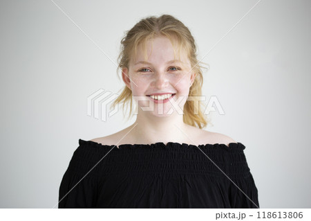 A happy young woman with blonde hair smiles warmly, showcasing her natural beauty in a simple black off-the-shoulder top. The soft, light backdrop enhances young adult cheerful disposition. 118613806