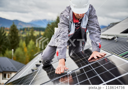 Man engineer mounting photovoltaic solar panels on roof of house. Technician in helmet installing solar module system with help of hex key. Concept of alternative, renewable energy. Man engineer mounting photovoltaic solar panels on roof of house. Technician in helmet installing solar module system with help of hex key. Concept of alternative, renewable energy. 118614736