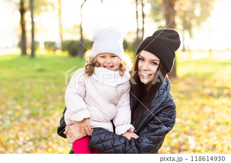 Mother and young little girl with blonde hair are playing in an autumn park on a yellow and orange leaf background 118614930