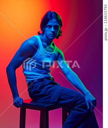 fashion portrait of young attractive man posing sitting on wooden stool dressed casual outfit in blue neon light against gradient studio background. fashion portrait of young attractive man posing sitting on wooden stool dressed casual outfit in blue neon light against gradient studio background. 118615793