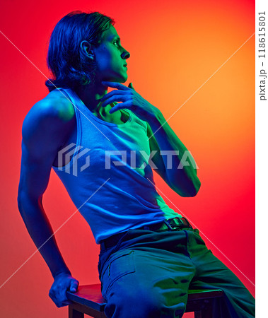 Stylish, attractive young man with athletic build body sitting on bar stool in blue neon light against gradient studio background. 118615801