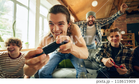 Intense focus on screen as player leans in with excitement, controller in hand, while his friend cheers him on in background. Intense focus on screen as player leans in with excitement, controller in hand, while his friend cheers him on in background. 118615909