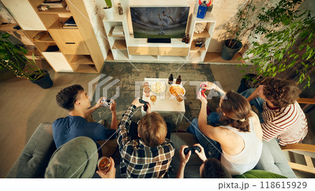 View from above of friends huddled around table, controllers gripped tightly, snacks within easy reach, while they battle on big screen. View from above of friends huddled around table, controllers gripped tightly, snacks within easy reach, while they battle on big screen. 118615929