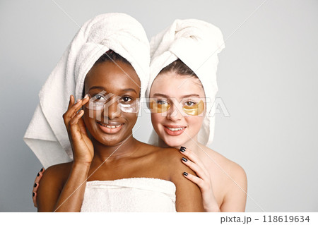Two young women in white bathrobes standing isolated on white. Girls wearing white towels on their hair. Girls have a eye patches on their faces. 118619634
