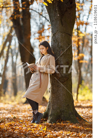 Beautiful woman reading a book in autumn park. Young girl standing near tree with a book. Brunette woman wearing beige coat. 118619681