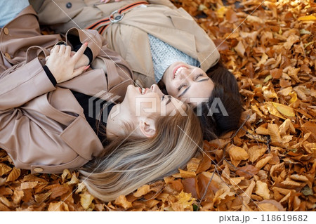 Two girls lying on the yellow leaves at the autumn park. Portrait of female friends. Two girls wearing beige coats. 118619682