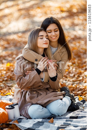 Two beautiful female friends spending time on a picnic blanket on the grass. Two young smiling sisters making picnic and hugging in autumn park. Brunette and blonde girls wearing coats. 118619683