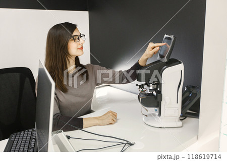 Female optician using laptop at her working place. Woman wearing eyeglasses in ophthalmic shop. Optician checking eyeglasses in special machine 118619714