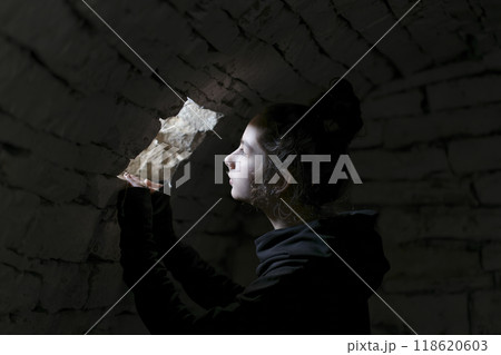 Young adult in a basement looking in a small window. A beam of light falls on her face. Portrait of the young girl in the dungeon with dark background. Young adult in a basement looking in a small window. A beam of light falls on her face. Portrait of the young girl in the dungeon with dark background. 118620603