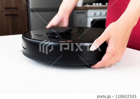 Black robot vacuum cleaner in female hands, close-up. The woman puts the vacuum cleaner on the table to disassemble and clean it after cleaning 118622583