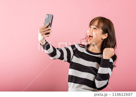 Happy Asian portrait beautiful cute young woman teen smiling excited using mobile phone say yes Studio shot isolated on pink background, Thai female surprised make winner gesture on smartphone 118622687