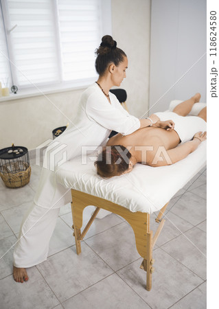 Process of back massage in pro salon. Caucasian female therapist at work with woman laying face down on massage table, vertical image, selective focus. Process of back massage in pro salon. Caucasian female therapist at work with woman laying face down on massage table, vertical image, selective focus. 118624580