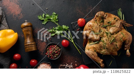 Baked chicken on a cutting board with rosemary, cherry tomatoes and spices. Dark background, wooden cutlery and kitchen utensils Baked chicken on a cutting board with rosemary, cherry tomatoes and spices. Dark background, wooden cutlery and kitchen utensils 118625822