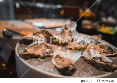 A bowl of fresh oysters on ice in an elegant white bowl, set against a blurred background of a luxury hotel's buffet, showcasing high-end dining and attention to detail. 118626530
