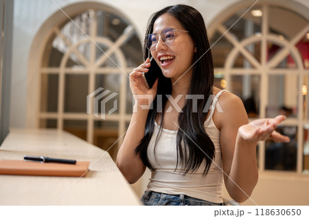 A beautiful, cheerful Asian woman is enjoying a phone conversation while relaxing in a coffee shop. 118630650