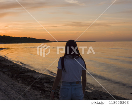 As the sun sets on the seashore a young woman stands in awe, her silhouette framed by the brilliant colors of the sunset, creating a serene moment of reflection and beauty. As the sun sets on the seashore a young woman stands in awe, her silhouette framed by the brilliant colors of the sunset, creating a serene moment of reflection and beauty. 118630868