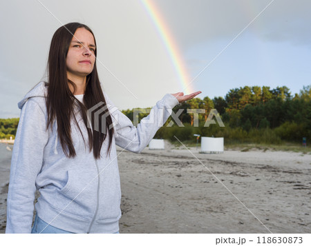 On the beach, Woman Young Brunette reaches out, catching a Rainbow in her hand. The delicate hues of the Rainbow dance against backdrop of waves, as she gazes at Rainbow's shimmering light. 118630873