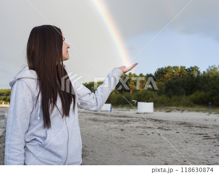 A Woman Young Brunette stands on the beach her hand gently cradling a vibrant Rainbow. The colors of Rainbow contrast beautifully with the sandy shore as the Rainbow arches gracefully into her palm. 118630874