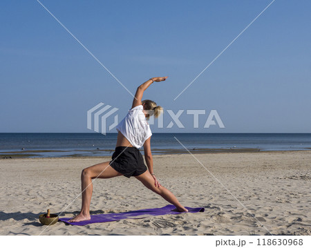 A young blonde woman stretches her muscles through yoga on a beach mat the sea as her backdrop. Her yoga practice flows smoothly, blending movement and mindfulness near the calming waves. A young blonde woman stretches her muscles through yoga on a beach mat the sea as her backdrop. Her yoga practice flows smoothly, blending movement and mindfulness near the calming waves. 118630968