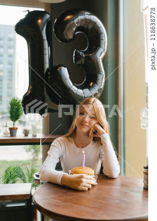 Happy teenage girl sits at a table with a birthday cake with lit candle and a balloon that says thirteen 118638778