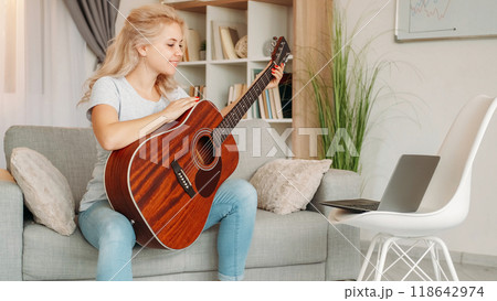 Online lesson. Happy woman. Instrumental playing. Excited lady holding acoustic guitar having video conference on laptop light room interior. 118642974