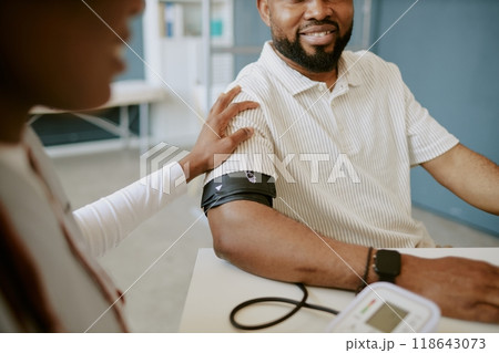 Smiling man sitting at medical examination table receiving blood pressure reading from a nurse in a clinical setting, with monitoring device and patient interaction present 118643073